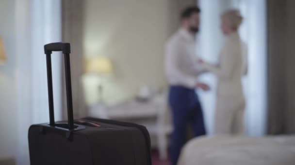 Close-up of travel bag with blurred adult man and woman hugging at the background. Young Caucasian couple of lovers meeting in hotel room. Love, leisure, travelling, joy, lifestyle. - Video