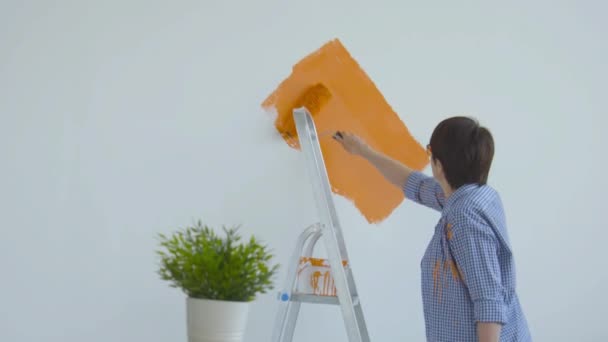 Σκέψη ανακαίνισης. Ευτυχισμένη μεσήλικη γυναίκα ζωγραφική λευκό τοίχο με ρολό χρώμα, πορτοκαλί χρώμα - Πλάνα, βίντεο
