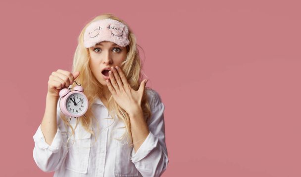 Slapeloze jonge vrouw in pyjama en slaapmaskers op een roze achtergrond. Alarm klok maakte het meisje wakker - Foto, afbeelding