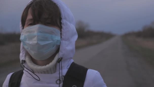 free girl traveler with a backpack in protective medical mask and hood is walking out of town on road. Protection against viruses and bacteria. health and safety concept, coronavirus N1H1, virus - Footage, Video