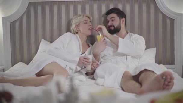 Smiling Caucasian man giving orange juice to girlfriend and grateful blond woman kissing lover on cheek. Happy couple resting in bed in luxurious hotel room. Love, togetherness, tourism, lifestyle. - Video