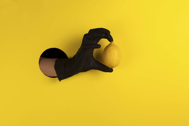 Hand in a black glove in a hole on a yellow background and offers lemon as a source of vitamin C and means of protection against coronavirus infection Covid-19. Copy space. - Фото, изображение