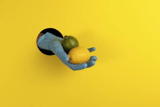 Medical concept. A hand in a blue glove in a hole on a yellow background and offers lemon and lime as a source of vitamin C & means of protection against coronavirus infection Covid-19. Copy space. - 写真・画像