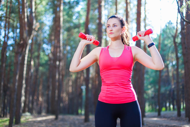 Sportive attractive girl working out with dumbbells, training in the forest. Fitness, sport, lifestyle concept - Foto, imagen