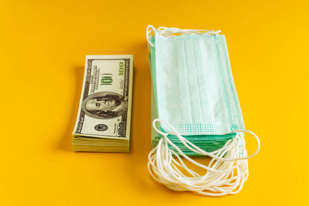 A stack of medical masks next to a stack of money on a yellow background. The concept of increasing the price of surgical masks due to the rapid spread of the coronavirus - Fotografie, Obrázek