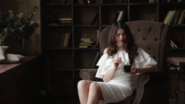 Young attractive woman wearing white dress is sitting in the great armchair and drinking coffee in the library - Imágenes, Vídeo