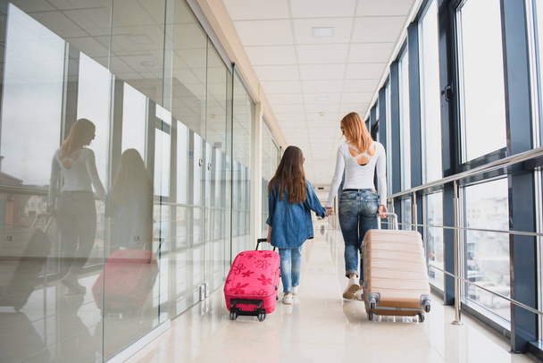 Happy young mother and her daughter walking in the airport terminal while carrying a suitcase. High season and vacation concept. Relax and lifestyles - Фото, изображение
