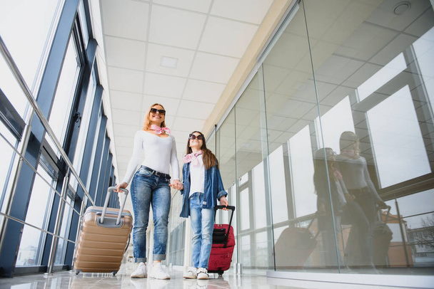 Beautiful mother and her stylish cute daughter walk together and pull a pink suitcase at airport. High season and vacation concept. Relax and lifestyles - Photo, Image