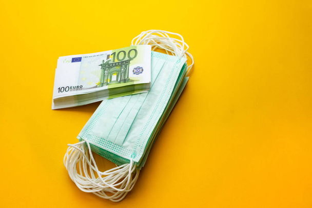 A stack of medical masks next to a stack of money on a yellow background. The concept of increasing the price of surgical masks due to the rapid spread of the coronavirus. - Foto, afbeelding