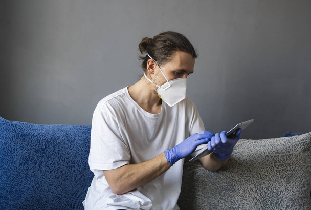 Man in white shirt, medical mask and rubber gloves sits at home and works with tablet on a sofa during quarantine. Man, designer, artist, architect, businessman at remote work in a pandemic covid - Zdjęcie, obraz