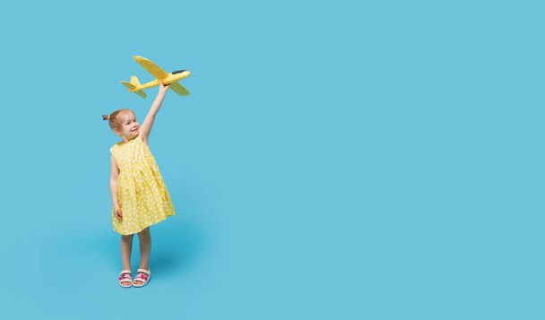 Κοριτσάκι με καλοκαιρινό φόρεμα κρατά στα χέρια του ένα αεροπλάνο-παιχνίδι σε μπλε φόντο. Ώρα για ταξίδι. - Φωτογραφία, εικόνα