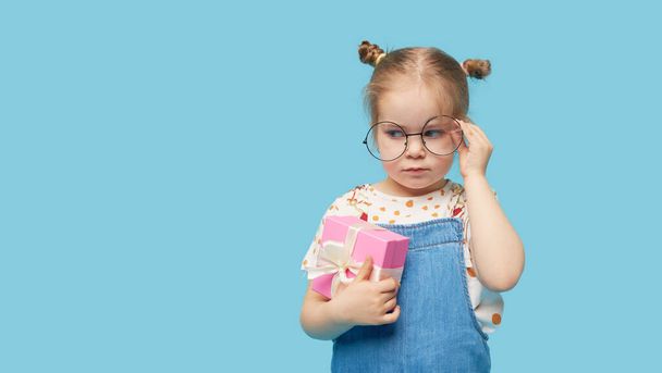 Portrait of upset, offended little toddler girl child standing isolated over blue background. Holding a gift box. Empty space for text - Foto, imagen