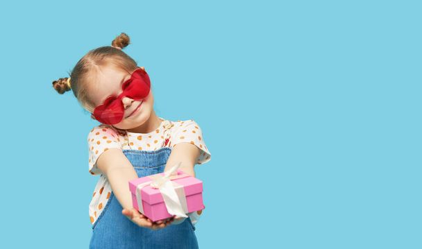 Portrait of surprised cute little toddler girl child standing isolated over blue background. Holding a gift box. Empty space for text - 写真・画像