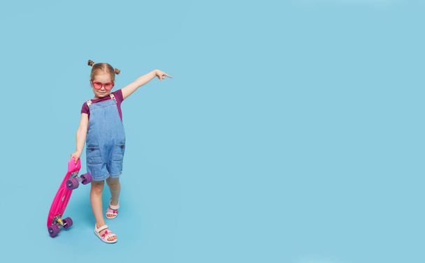 Cool little toddler girl in bright clothes sits on a skateboard and looking at the camera isolated on blue background - Zdjęcie, obraz