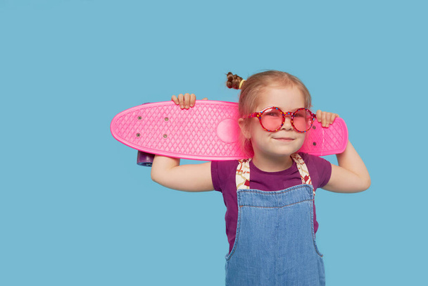 Cool malé batole dívka v jasném oblečení sedí na skateboardu a dívá se na kameru izolované na modrém pozadí - Fotografie, Obrázek