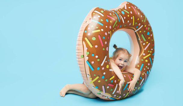 Failed summer vacation. Happy surprised toddler girl resting on a donut-shaped rubber ring. Sea vacation concept on a colored blue background. Wow emotions	 - Foto, Imagem