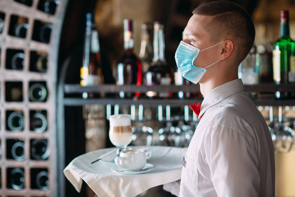 A European-looking waiter in a medical mask serves Latte coffee - Фото, изображение