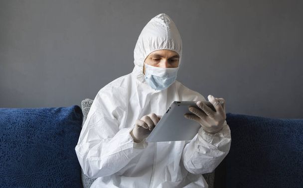 Man in protective suit, medical mask and rubber gloves sits at home and works with tablet on a sofa during quarantine. Designer, artist, architect, businessman at remote work in a pandemic covid. - Fotoğraf, Görsel