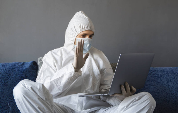 Man in protective suit, medical mask and rubber gloves sits at home and works with laptop at the table during quarantine. Designer, artist, architect, businessman at remote work in a pandemic covid. - Foto, Imagen