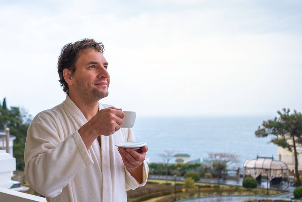 Happy man in a white bathrobe meets the morning with a cup of tea or coffee on a background of the sea. The concept of rest, health and awakening - Photo, image