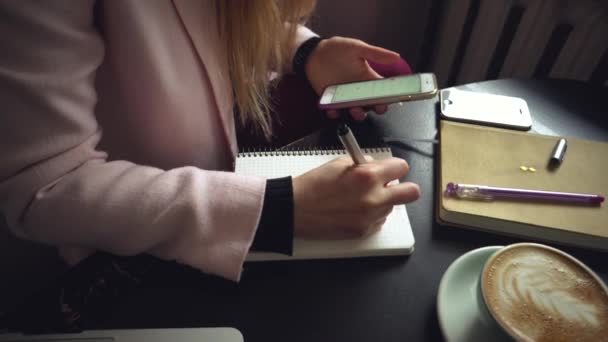 Lähikuva valkoihoinen nainen käsi puinen pöytä sisällä kahvila tekee muistiinpanoja muistikirjaan. Aiheena freelancer bloggaaja toimittaja työssä. Tunnistamaton henkilö. Teknologia puhelin ja kannettava tietokone kuppi kahvia - Materiaali, video