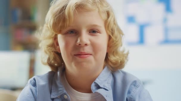 Portrét roztomilého malého chlapce s kudrnatými blond vlasy sedí u svého školního stolu, šťastně se usmívá. Chytrý chlapec s okouzlujícím úsměvem sedí ve třídě. Detailní záběr kamery - Záběry, video