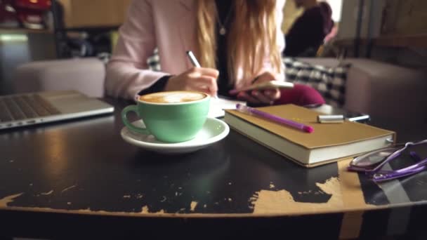 Feche a mão de mulher caucasiana na mesa de madeira dentro do café faz notas no caderno. Assunto jornalista blogueiro freelancer no trabalho. Pessoa irreconhecível. Tecnologia telefone e laptop com xícara de café
 - Filmagem, Vídeo