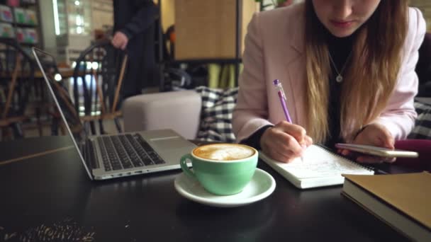 Close up caucasian woman hand on wooden table inside cafe makes notes in notebook. Subject freelancer blogger journalist at work. Unrecognizable person. Technology phone and laptop with cup coffee - Footage, Video