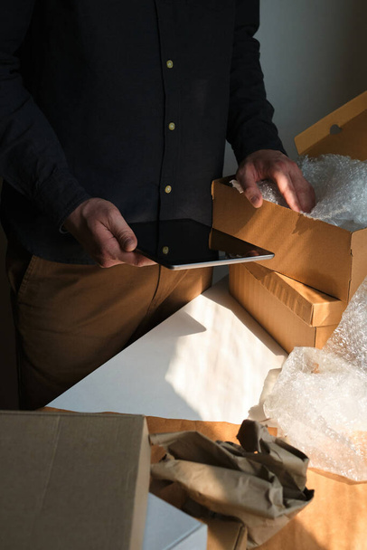 A man is packing cardboard boxes. Delivery service for online orders. Holding tablet in hand. The guy working in office, the cargo business. Warehouse with products, preparation for express shipping - Photo, image