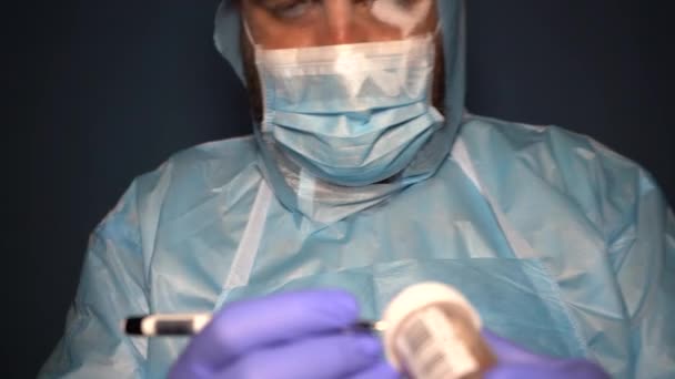 Ein bärtiger Arzt im Schutzanzug überprüft Laborflaschen zur Analyse während der Covid-19-Coronavirus-Pandemie. Müder Arzt in nebliger Maske. - Filmmaterial, Video