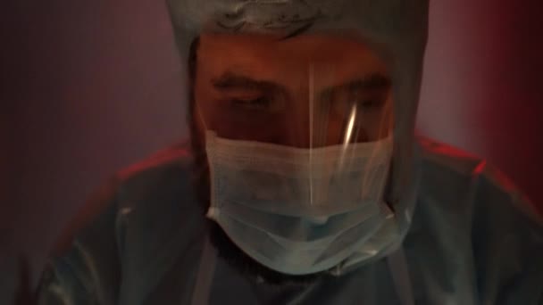 Een arts in een beschermend pak in een laboratorium met rood-blauw noodlicht. Laboratoriumnoodsituatie tijdens de coronaviruspandemie. - Video