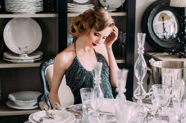 Beautiful blond girl with vintage make up and hairstyle in evening shining dress sitting at table with plates and glasses and looking to the side and hand close to head - Φωτογραφία, εικόνα