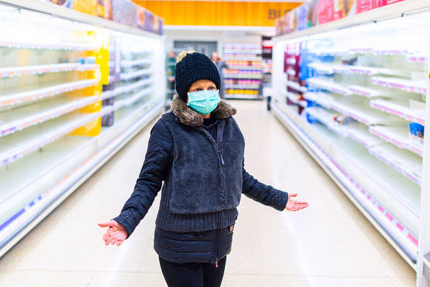 La mujer con máscara parece confundida en el pasillo entre estantes vacíos en un supermercado. Mascarilla protectora para la propagación de la enfermedad coronavirus. Epidemia de Coronavirus en la ciudad. COVID-19 coronavirus pandémico
. - Foto, imagen