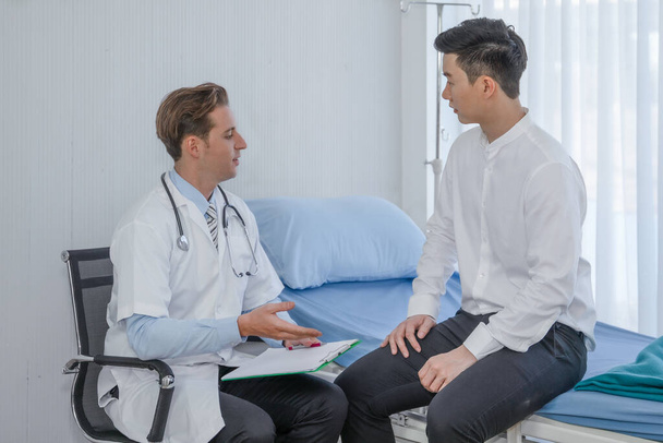 antecedentes de salud de caucásico médico masculino examinando paciente masculino asiático sentado en la cama en el hospital
 - Foto, Imagen