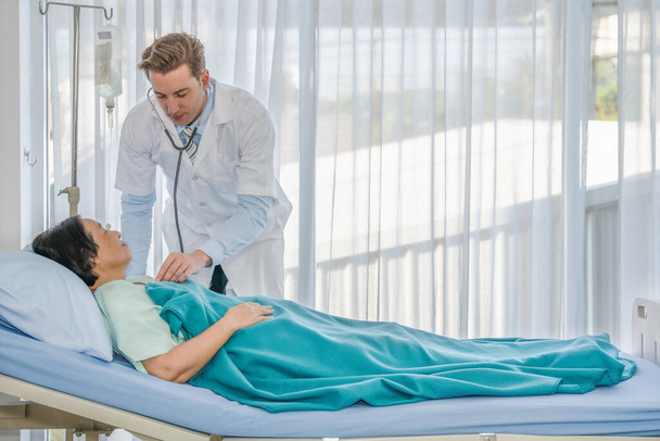 healthcare background of caucasian male medical doctor examining asian female senior patient lying on bed with stethoscope in hospital - Valokuva, kuva