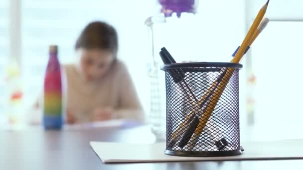little Israeli caucasian teen gifted girl taking a test in an elementary school class. She tries to solve the exam and writing her answers on paper. Closeup on a box with pens and pencils with blur BG - Footage, Video
