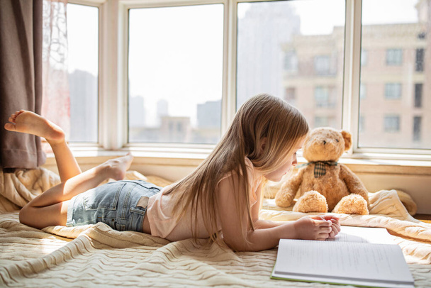 Roztomilá malá běloška v neformálním oblečení, čte si knížku s plyšovým medvídkem a usmívá se, zatímco leží na podlaze u okna v místnosti. Zůstaňte doma během pandemie coronavirus covid-19. - Fotografie, Obrázek