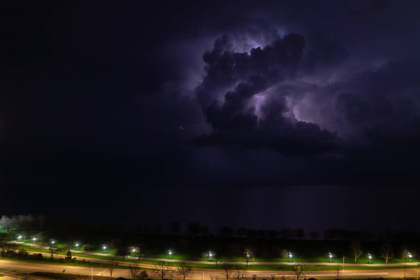 Интенсивное облако грозы синего или фиолетового цвета и освещаемое многочисленными ударами молнии движется над озером Мичиган, когда машины проходят по Лейк Шор Драйв на северной стороне Чикаго
. - Фото, изображение