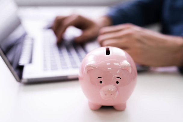 Close-up Of Businessman's Hand Working On Laptop With Pink Piggybank - 写真・画像