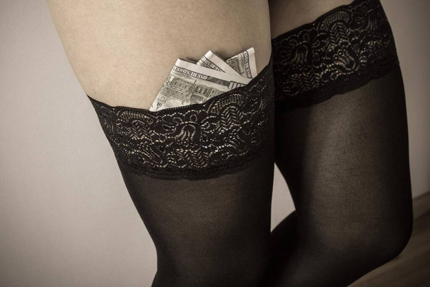 In stockings shoved money. Venal sex, love, payment services. Silence fee. Female legs, light background. Muffled tones, vignetting. - Foto, afbeelding