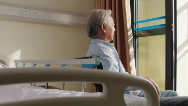 Ασιάτης γέρος κάθεται σε αναπηρική καρέκλα στο δωμάτιό του στο γηροκομείο και φαίνεται χαρούμενος και ικανοποιημένος - Πλάνα, βίντεο