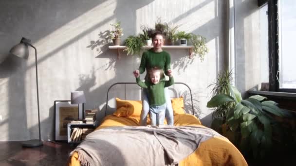Mom and daughter dressed equally in green sweaters and jeans are jumping on the bed. Slow motion video - Footage, Video