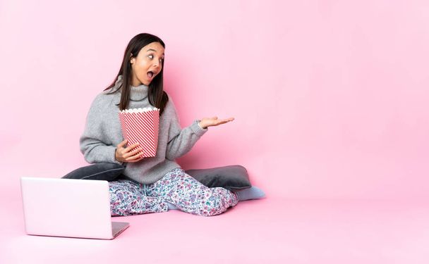 Young mixed race woman eating popcorn while watching a movie on the laptop presenting an idea while looking smiling towards - Photo, Image