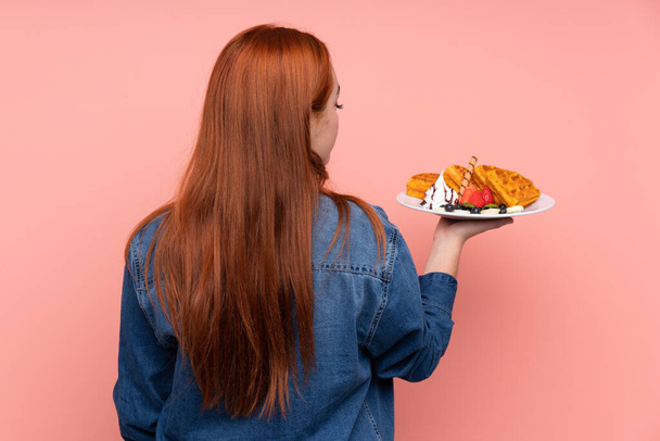 Redhead teenager dívka drží vafle přes izolované růžové pozadí v zadní poloze - Fotografie, Obrázek