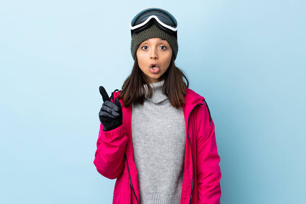 Змішана гоночна лижниця дівчина зі сноубордистськими окулярами над ізольованим синім тлом думає про ідею, спрямовану на палець вгору
. - Фото, зображення