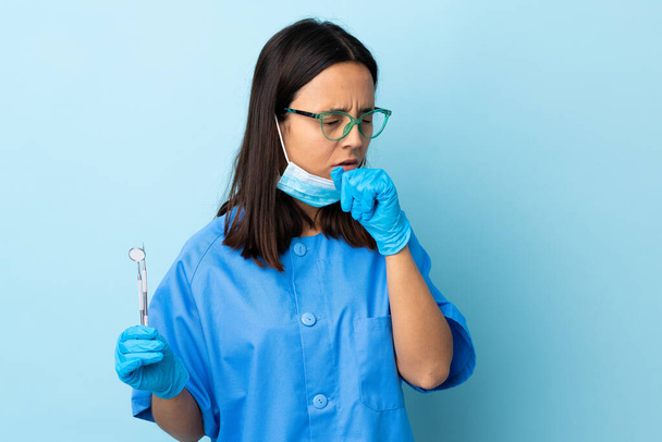 Молода брюнетка змішаної раси стоматолог жінка тримає інструменти над ізольованим фоном кашляє багато
 - Фото, зображення