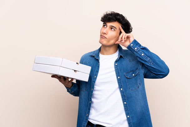 Mladý argentinský muž drží pizzy přes izolovanou zeď s pochybnostmi a zmateným výrazem ve tváři - Fotografie, Obrázek