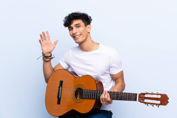 Giovane uomo argentino con chitarra su sfondo blu isolato che saluta con mano con espressione felice
 - Foto, immagini