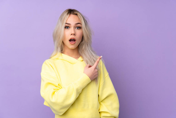 tiener meisje het dragen van een gele sweater over geïsoleerde paarse achtergrond verrast en wijzen kant - Foto, afbeelding
