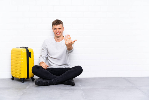 Young handsome man sitting on the floor with a suitcase inviting to come - Foto, Imagem